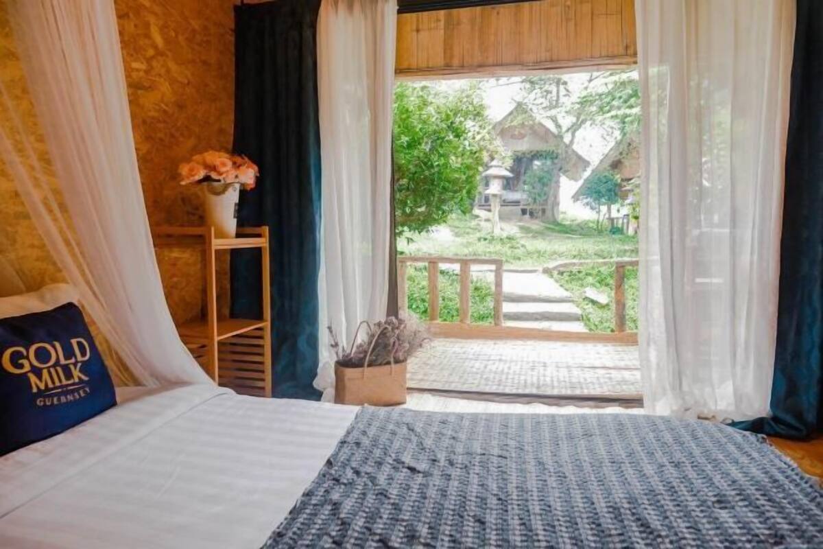 A bedroom with a bed, a nightstand, a vase with flowers, and a view of a garden and house through a window.