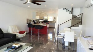 Un moderno salón con un sofá negro, una mesa de centro de vidrio y un sillón rojo. Hay una zona de cocina con una isla roja y taburetes de bar. Una escalera con barandilla de madera conduce a un nivel superior.