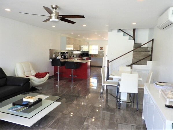 Un moderno salón con un sofá negro, una mesa de centro de vidrio y un sillón rojo. Hay una zona de cocina con una isla roja y taburetes de bar. Una escalera con barandilla de madera conduce a un nivel superior.