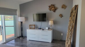 Una habitación moderna con una televisión grande, un tocador blanco, dos lámparas y vistas a una masa de agua a través de una puerta corrediza.
