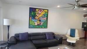 Ein Wohnzimmer mit einem schwarzen Ledersofa, einem weißen Sessel, einer Stehlampe und einem farbenfrohen, abstrakten Gemälde an der Wand.