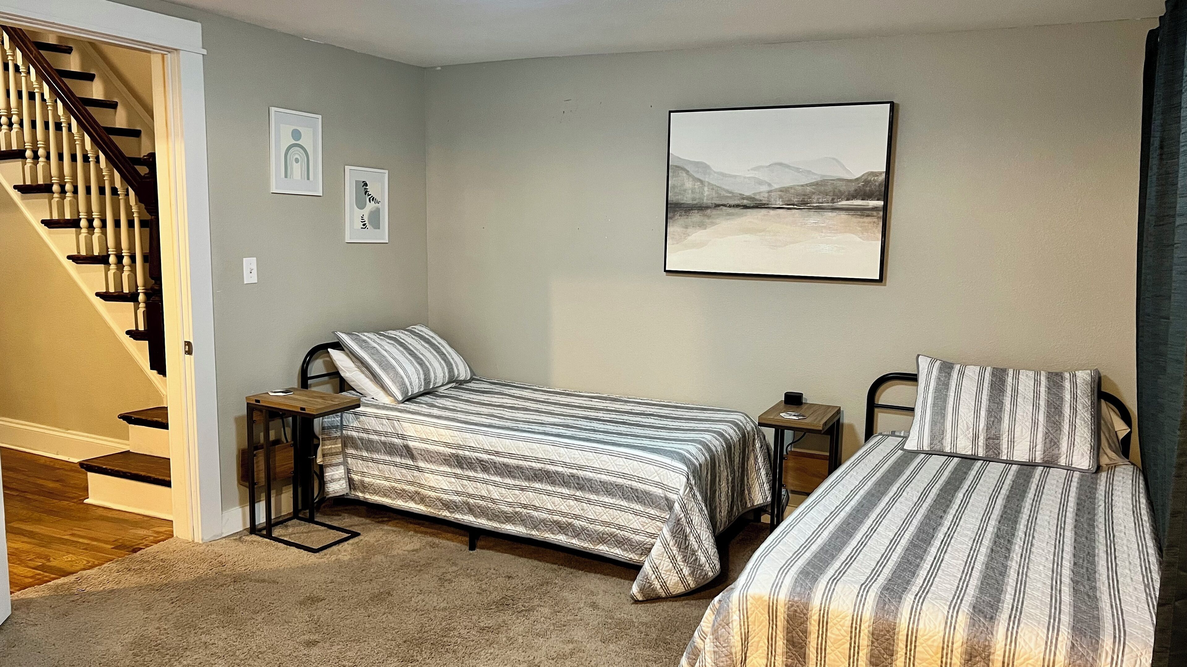 A bedroom with two single beds, a staircase, and a framed landscape painting on the wall.