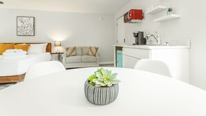 Ein modernes Hotelzimmer mit Bett, Sofa, Esstisch mit Topfpflanze, einer Küchenzeile mit roter Mikrowelle und einer kleinen Pflanze auf einem Regal.