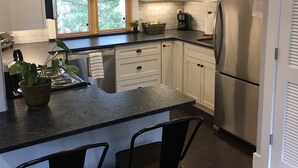A modern kitchen with white cabinets, a stainless steel refrigerator, a large window with blinds, and a small island with bar stools.