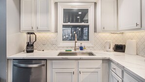 A modern kitchen with white cabinets, a stainless steel dishwasher, a built-in window, and a countertop with a blender and toaster. -  Modern 4BR Near Downtown, Chef Kitchen, Fenced Yard, EV Charger, W/D (Omaha)
