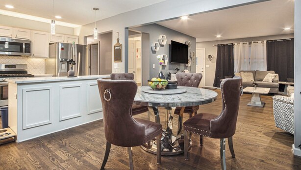 A modern kitchen with white cabinetry, a central island, and stainless steel appliances. There is a dining area with a round table and brown leather chairs. -  Modern 4BR Near Downtown, Chef Kitchen, Fenced Yard, EV Charger, W/D (Omaha)