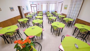 A dining area with tables and chairs arranged in rows, a potted plant with orange flowers, and a door with a sign above it. - Pastor Angelicus - Casa per Ferie (Chiusi della Verna)