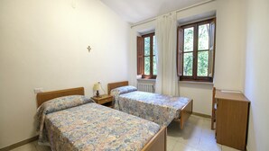 A bedroom with two beds, a nightstand, a lamp, a window with curtains, and a radiator. - Pastor Angelicus - Casa per Ferie (Chiusi della Verna)