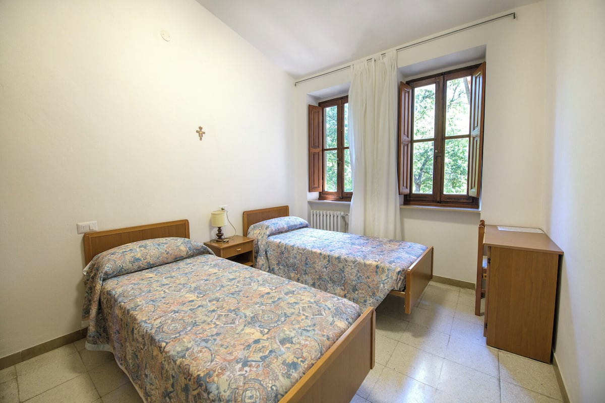 A bedroom with two beds, a nightstand, a lamp, a window with curtains, and a radiator.