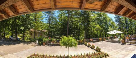 Un espacio al aire libre techado, con techo de madera, rodeado de árboles y un patio empedrado con bancos y jardineras.