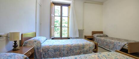 Quarto com três camas, janela com cortinas, radiador e abajur sobre a mesa de cabeceira.