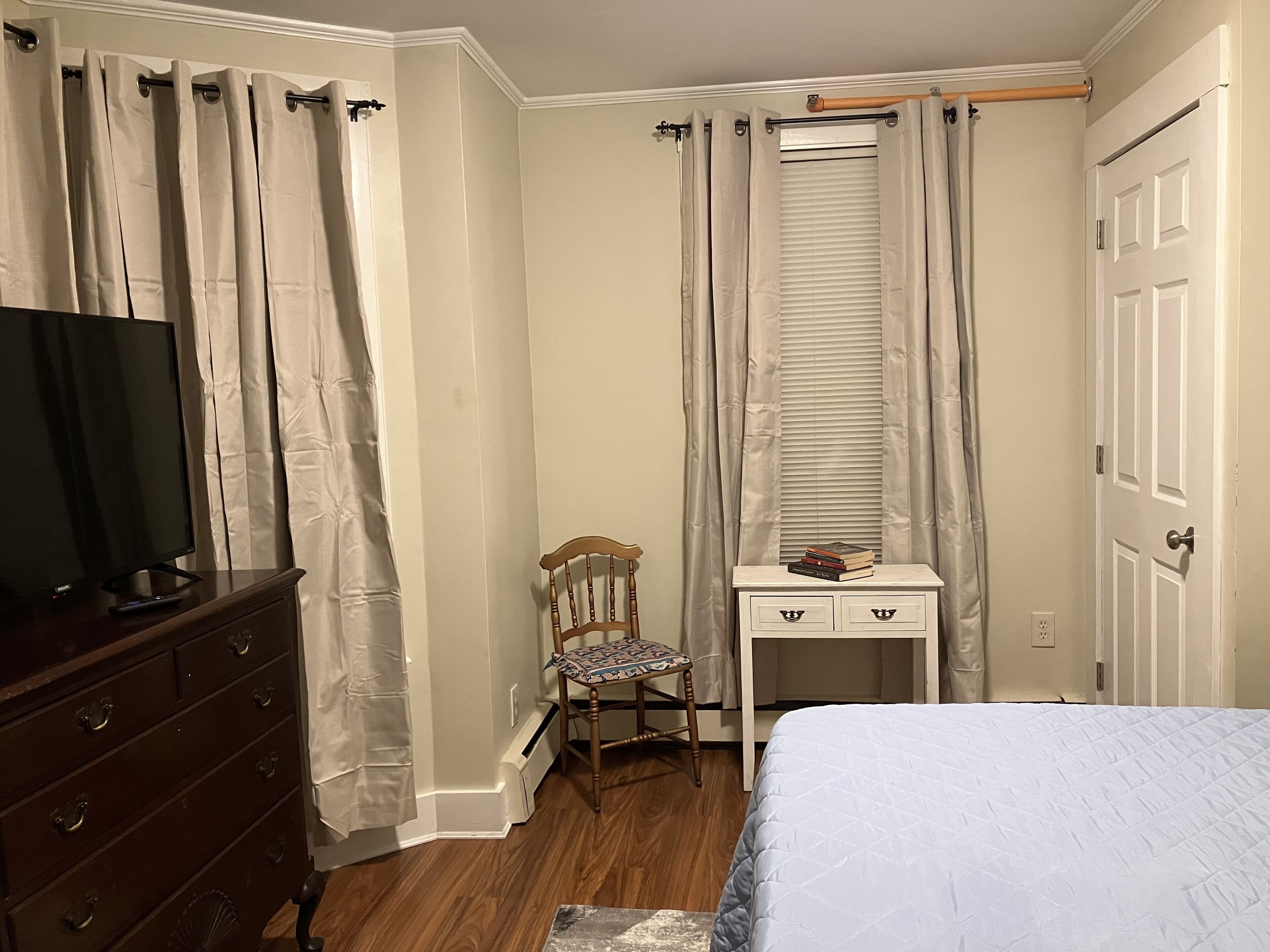 Ein Schlafzimmer mit einem Bett, einem Schminktisch, einem Sessel, einem Schreibtisch mit Büchern und einem Fenster mit Vorhängen.