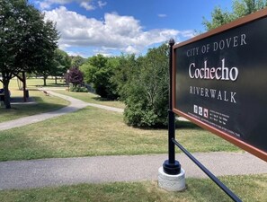 A sign for the City of Dover, Cochecho Riverwalk with a paved walkway and greenery.