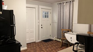Eine Küche mit Kühlschrank, Herd und Kaffeemaschine. Eine Tür führt in einen anderen Raum, und es gibt ein Fenster mit Vorhängen.