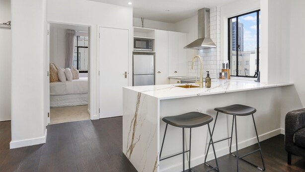 A modern kitchen with a white marble island, two bar stools, a stainless steel range hood, and a large window offering a view of the city. - Elegant Living in the City (Auckland)