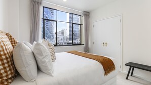 A hotel room with a large bed, a window with a city view, a bench, and a door. - Elegant Living in the City (Auckland)