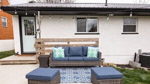 Een terras met een rieten bank en twee voetenbankjes, een kamerkleed met dessin, en een klein houten bankje.