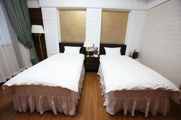 A hotel room with two single beds, a nightstand with a lamp, and a window with blinds. - Happiness CC Hotel (Naju)