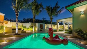 Une piscine agrémentée d’un flotteur en forme de flamant rose, entourée de palmiers et dotée d’un patio couvert.