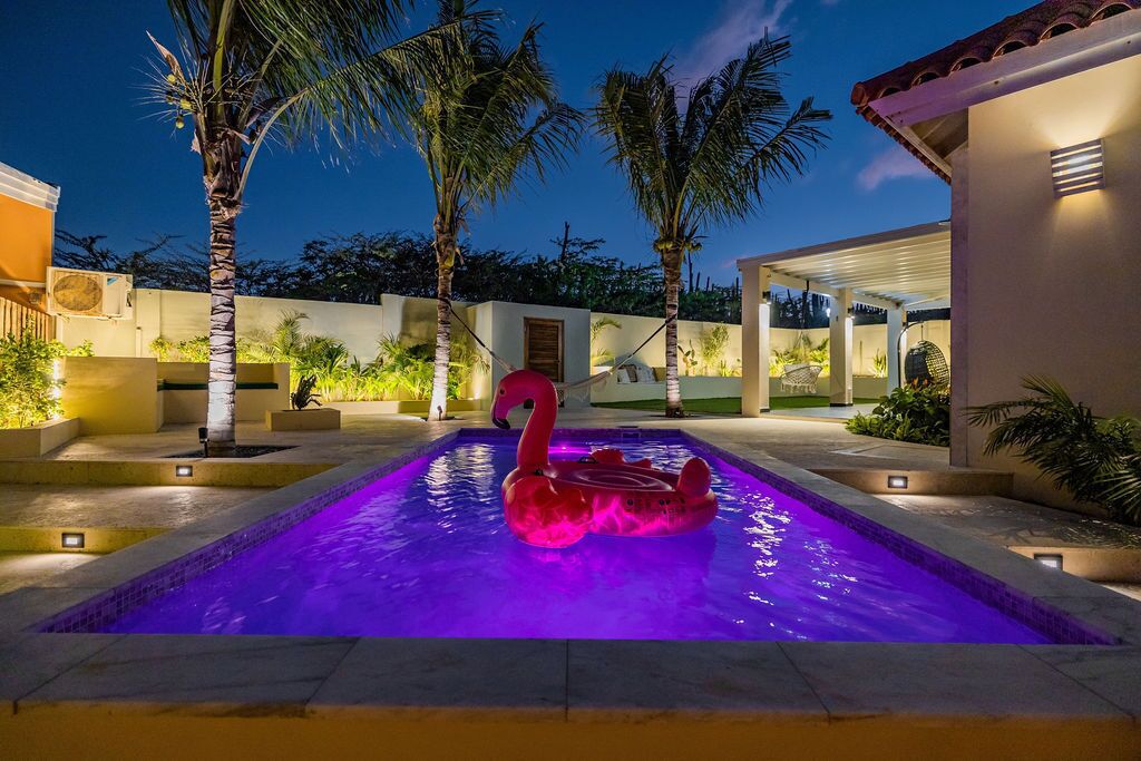 En swimmingpool med en flamingo-svømmebøje, omgivet af palmer og en overdækket terrasse.