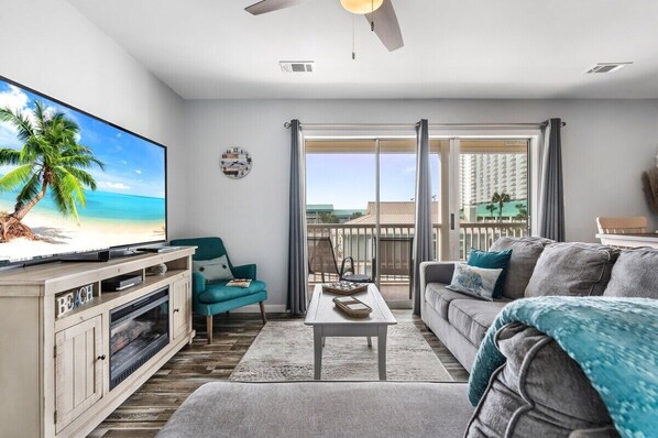 A living room with a TV, a sofa, a coffee table, and a balcony with a view of buildings.
