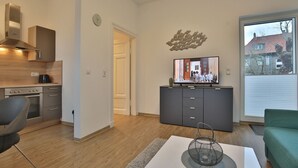 Eine moderne Küche mit Herd, Backofen und integrierter Dunstabzugshaube. Ein Essbereich mit Tisch und Stühlen, ein großes Fenster mit Blick auf Gebäude und ein Flachbildfernseher auf einem Schrank.