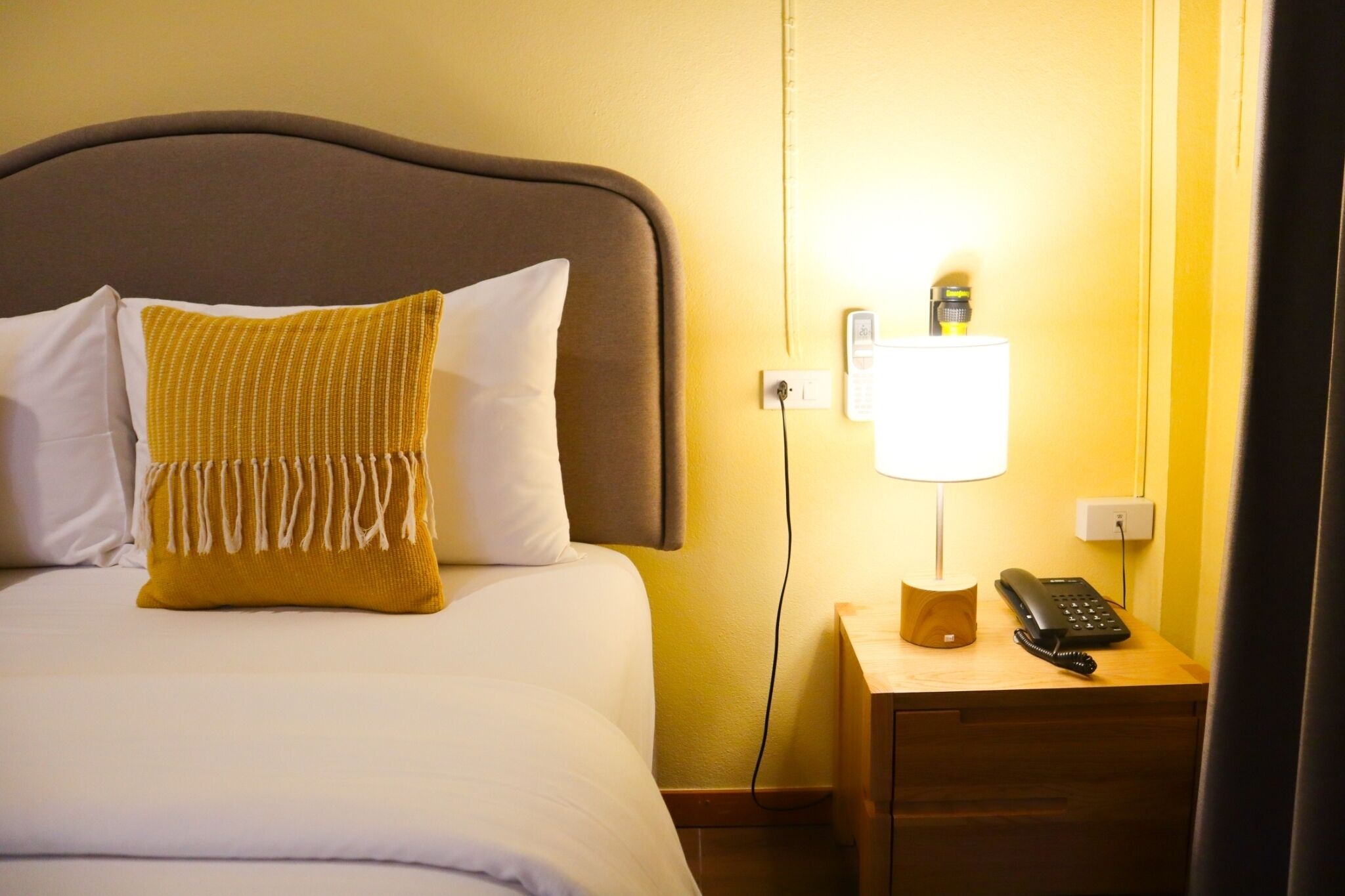 Une chambre d’hôtel comprenant un lit, une table de chevet avec une lampe et un téléphone, et une prise électrique au mur.