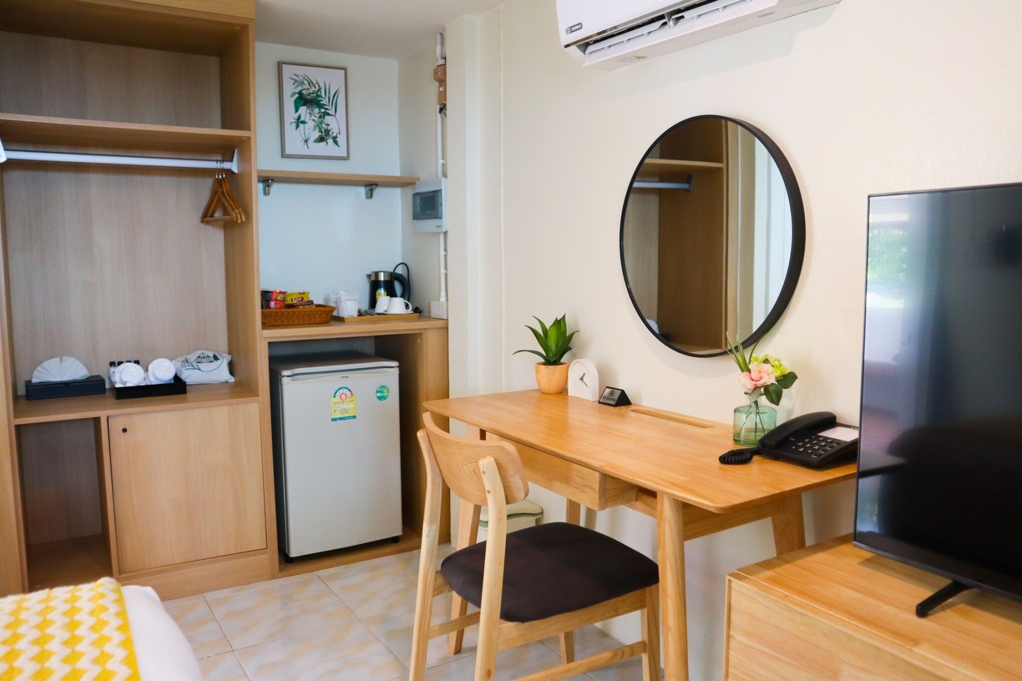 A compact hotel room with a wooden desk, a small refrigerator, a TV, a mirror, and a bed with a yellow and white patterned bedspread.