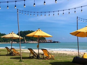 Ein Strand mit gelben Sonnenschirmen und Liegestühlen, einer Lichterkette und Blick auf das Meer und das entfernte Land.