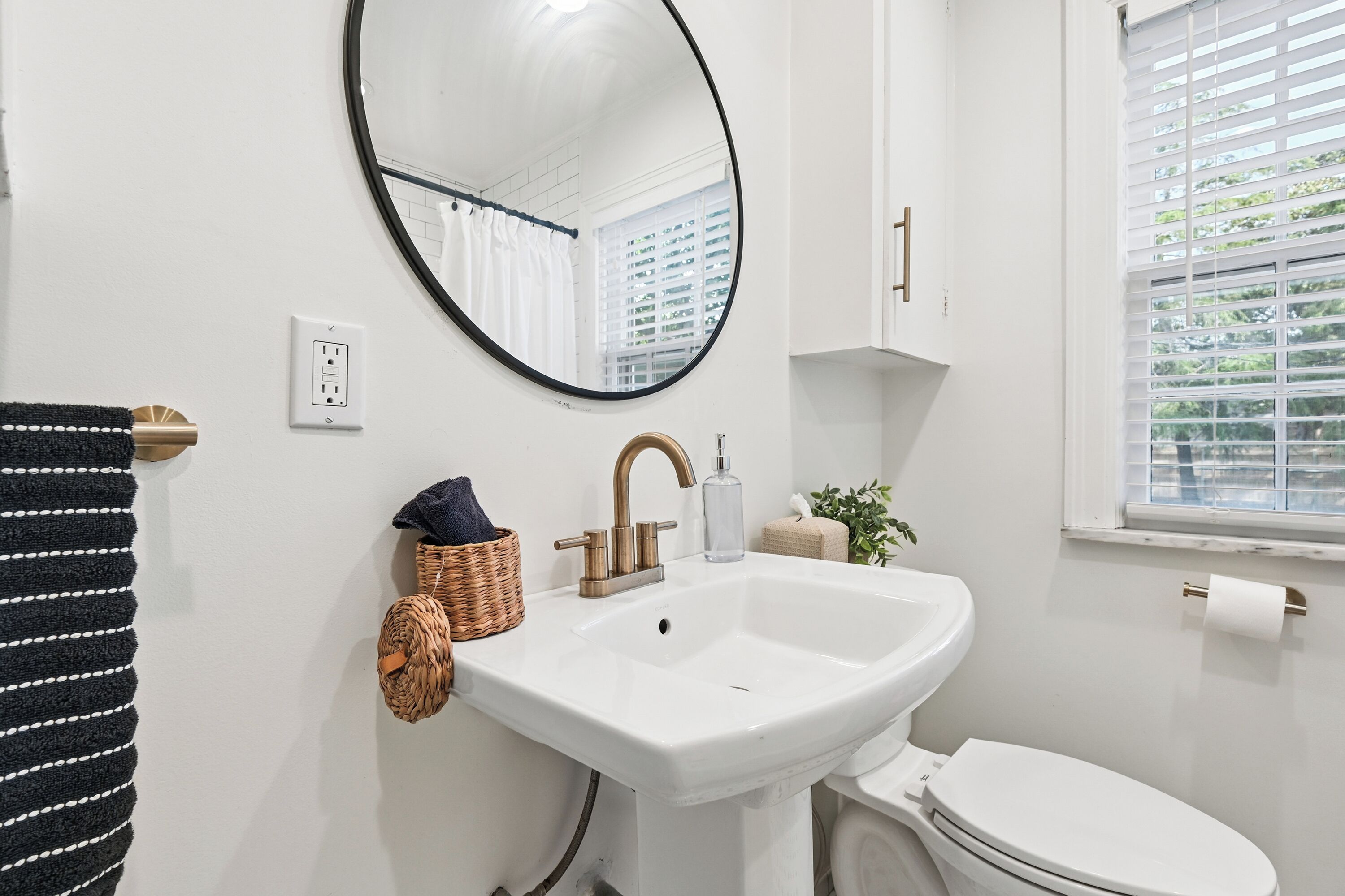 A bathroom with a white sink, a round mirror, a white toilet, a window with blinds, and a towel rack.