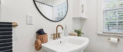 A bathroom with a white sink, a round mirror, a white toilet, a window with blinds, and a towel rack.