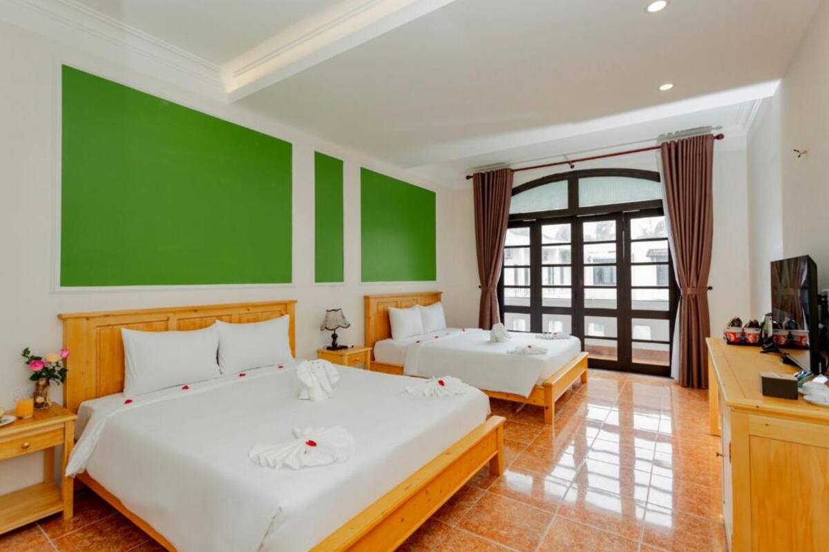 A hotel room with two beds, a large window with curtains, a wooden desk, and a green accent wall.