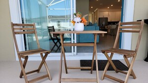 En uteplats med två trästolar och ett bord, en vas med blommor och en utsikt över interiören genom glasdörrar.