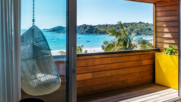 A balcony with a hanging chair, wooden floor, and a view of the sea with boats. - Oneroa Beach Apartment | You've Got It Maid (Waiheke Island)