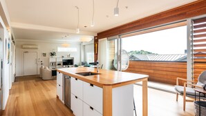 A modern kitchen with a wooden island, white cabinets, and a large window with blinds. - Oneroa Beach Apartment | You've Got It Maid (Waiheke Island)