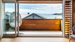 A modern interior with a sliding glass door leading to a balcony overlooking a body of water and distant hills. - Oneroa Beach Apartment | You've Got It Maid (Waiheke Island)