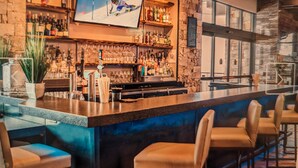 Uma área de bar com um balcão comprido, banquetas, estantes com garrafas e uma tela de televisão exibindo um esquiador.