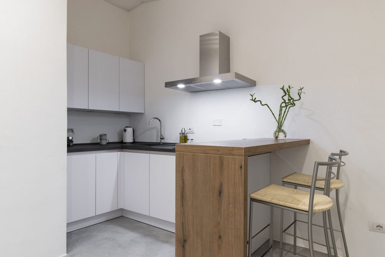 Una cucina compatta con mobili bianchi, una cappa in acciaio inossidabile, un piano di lavoro in legno e due sgabelli da bar.