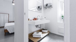 Casa de banho moderna com lavatório branco, espelho e porta-toalhas. Tem uma prateleira de madeira com toalhas e uma cesta.