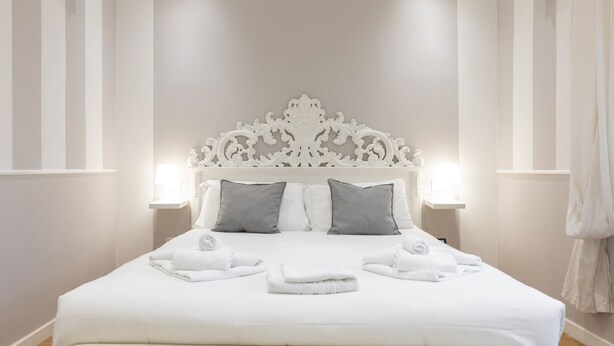 Cama bem arrumada com lençóis brancos e duas almofadas, cabeceira branca decorativa e duas luminárias de cabeceira.