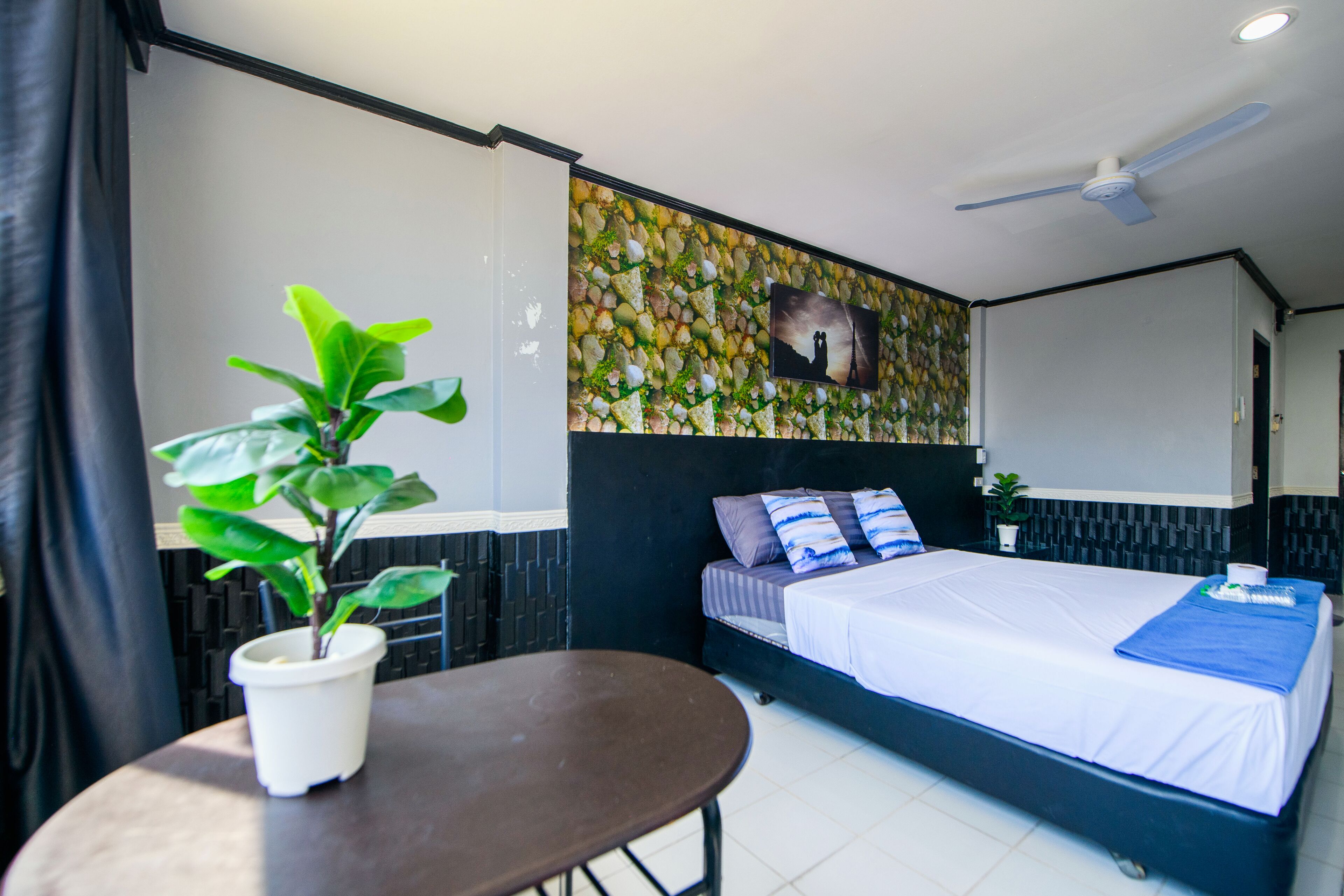 A bedroom with a bed, a ceiling fan, a potted plant, and a wall with a decorative pattern.