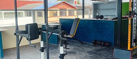 Un espacio de gimnasio al aire libre cubierto, con equipamiento de ejercicio, un banco y un soporte para pesas.