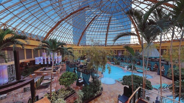 Un área de piscina interior con techo de cristal, sillones de descanso y palmeras.