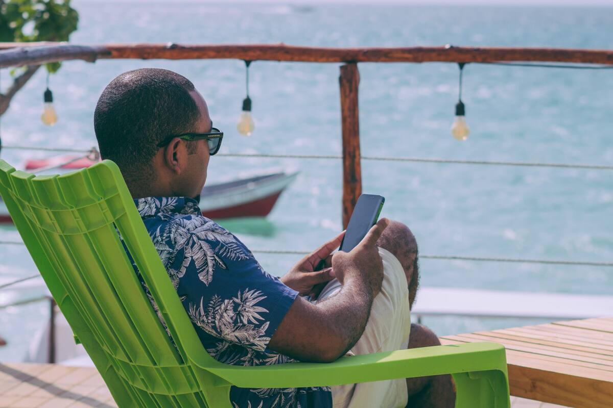 Un hombre sentado en una silla verde junto al mar, usando un teléfono inteligente.