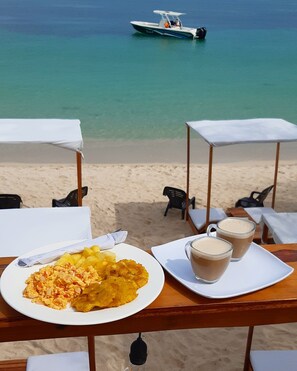 Ein Tisch am Strand mit Essen, zwei Gläsern und einem Boot im Hintergrund.