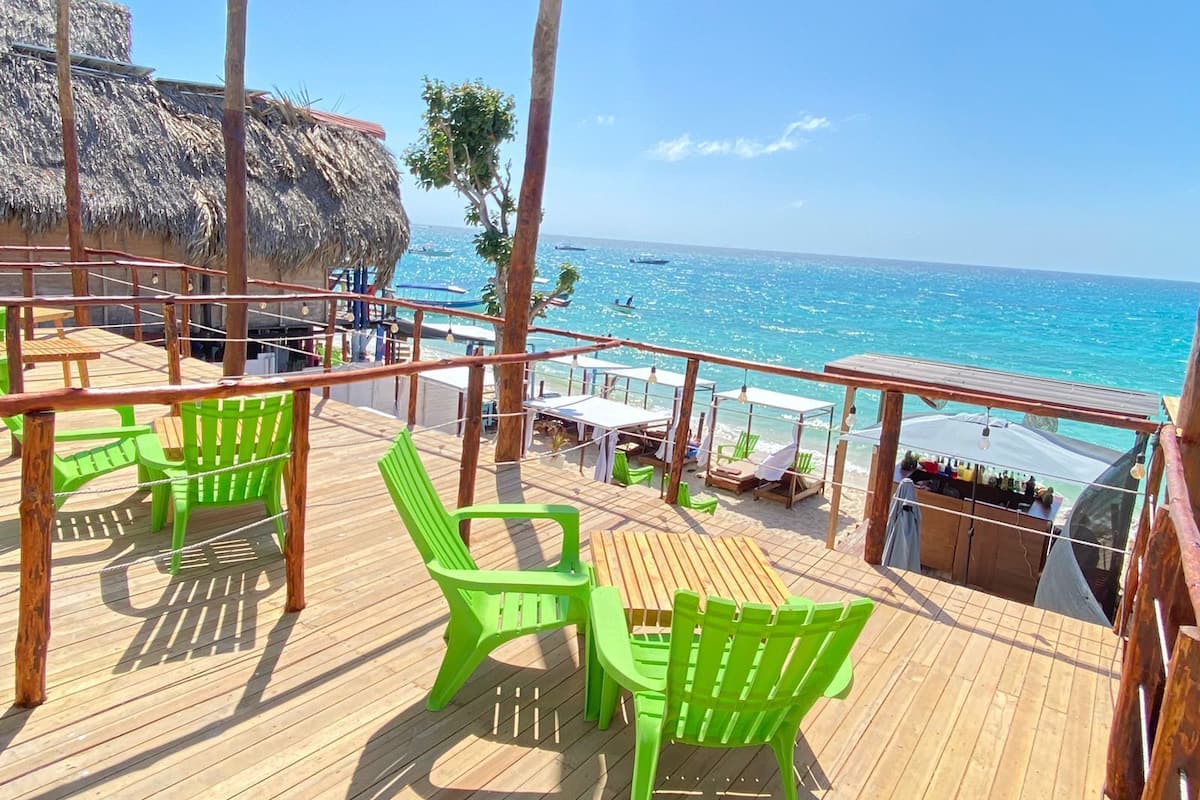 Una terraza de madera con sillas y mesas verdes, con vista a una playa y una estructura de techo de paja.