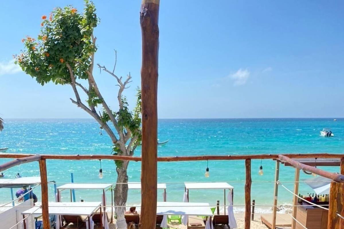Una terraza de madera con dos sillas de plástico verde y una mesa, con vista a una playa de aguas turquesas cristalinas y un barco a lo lejos.