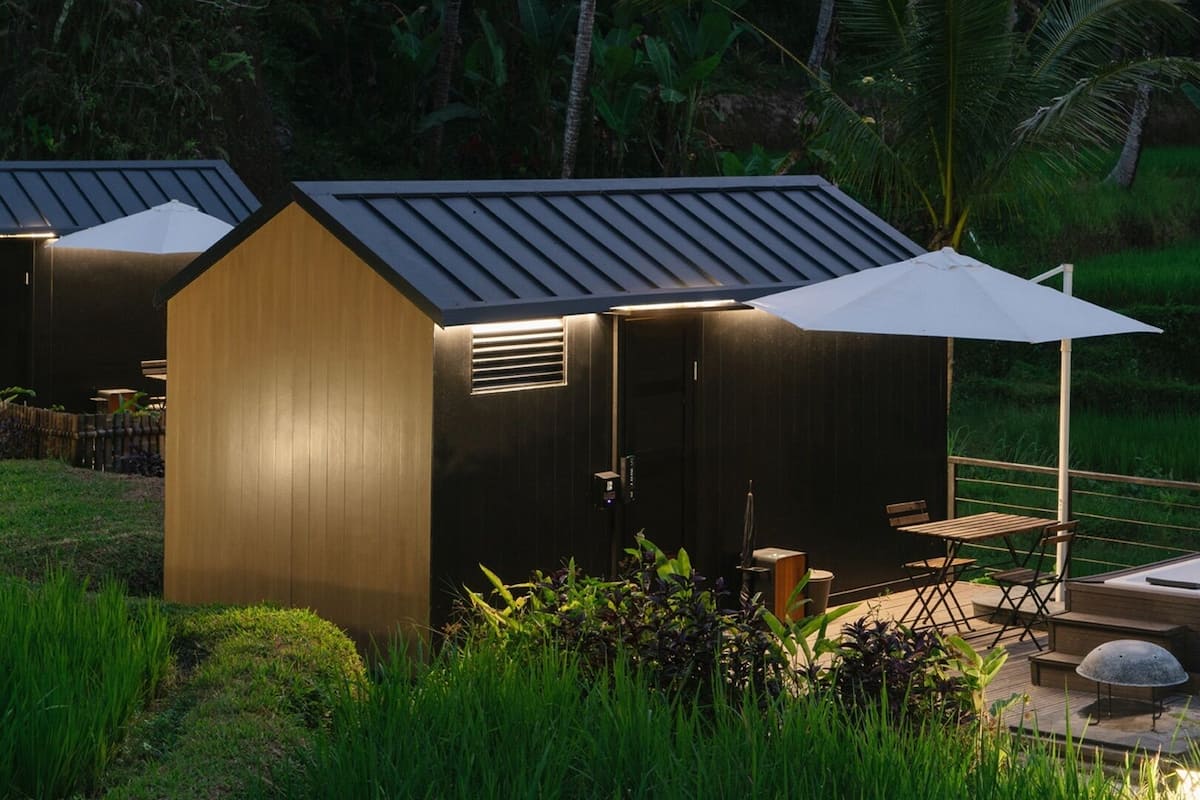 Una cabaña pequeña y moderna con techo inclinado y un paraguas grande que da sombra. Cuenta con una terraza de madera con asientos y un pequeño hogar.