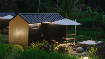 Una cabaña pequeña y moderna con techo inclinado y un paraguas grande que da sombra. Cuenta con una terraza de madera con asientos y un pequeño hogar.