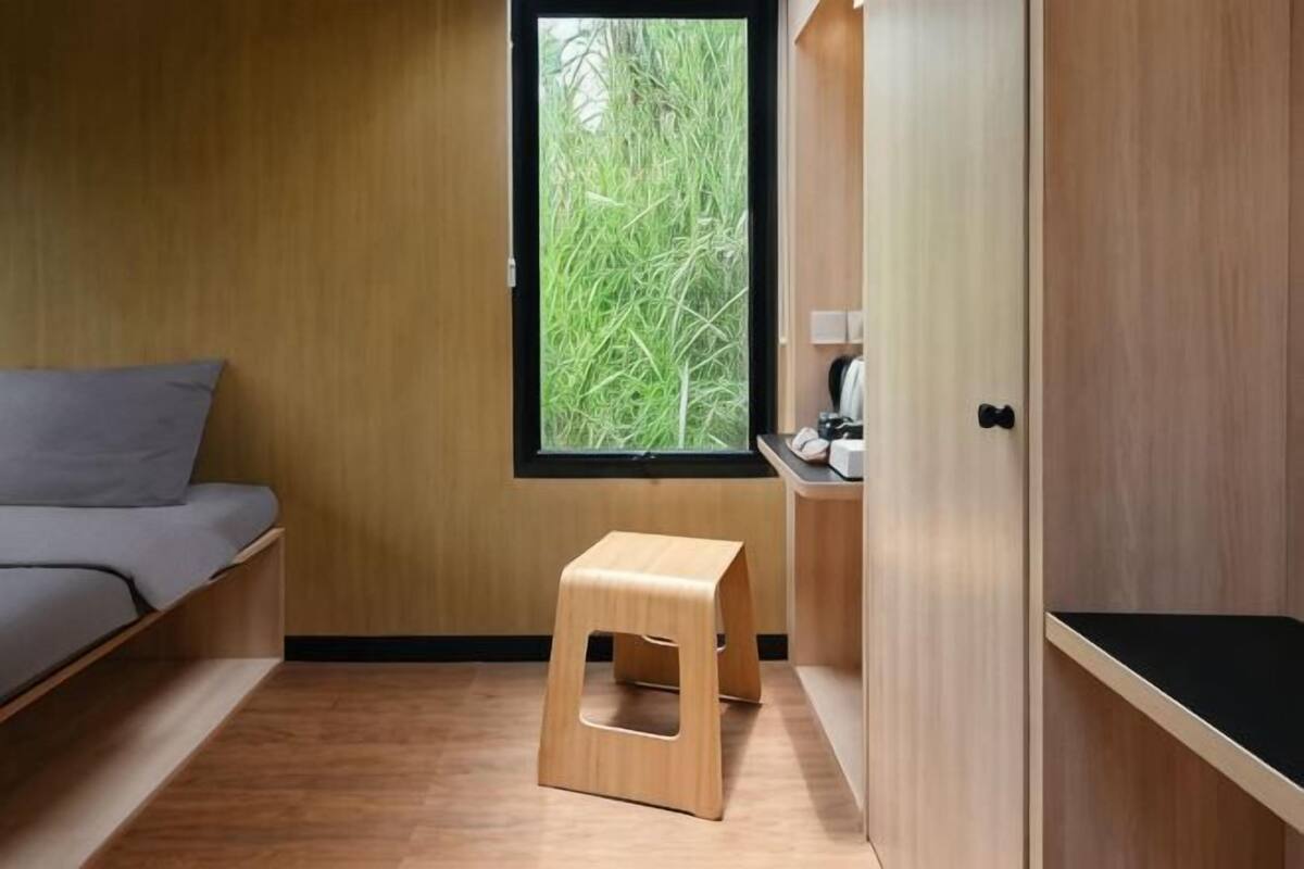 Una habitación con cama, un taburete, una ventana con vistas a la vegetación y un armario.